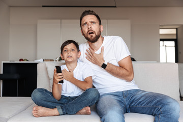 Image of outraged emotional man 30s with son 8-10 watching tv, while sitting on sofa in room