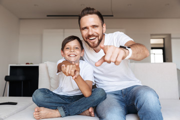 Image of cheerful glad man 30s and boy 8-10 laughing, while pointing finger at you and sitting on sofa in apartment