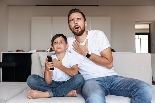Image of outraged emotional man 30s with son 8-10 watching tv, while sitting on sofa in room