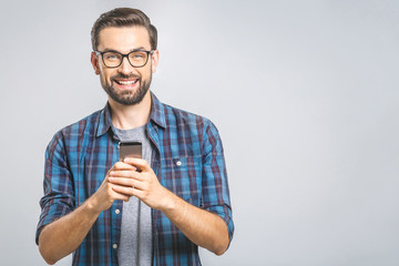 Happy young man in glasses typing sms on gray background, smiling and looking at camera.