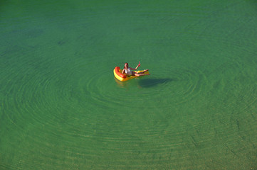 Fototapeta premium A young woman spends leisure time on an inflatable sun bed in the middle of a green lake. She wears a white swim suit and enjoys the summer sun as she relaxes on the sun bed. 