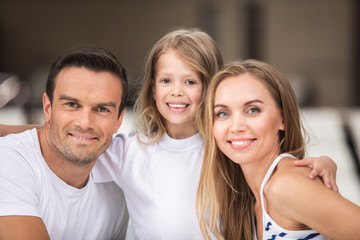 Happy together. Waist up portrait of smiling parents bonding to their kid. Small child is standing and embracing mother and father with content while they are sitting and looking straight with delight