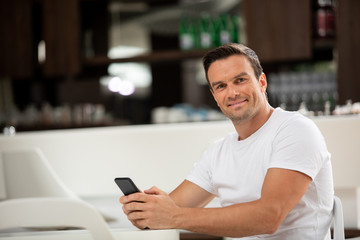 Waist up portrait of joyful man sitting at table in cafe. He is holding his phone in hands and surfing net with smile
