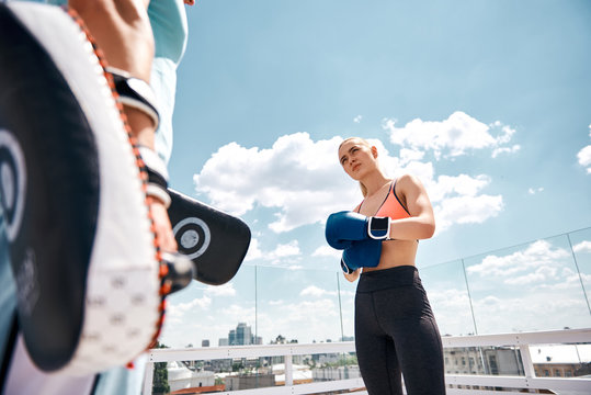 Low angle of fit woman during boxing training. She is standing and looking to her male instructor during short rest. They are exercising on open terrace of high urban house