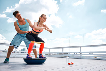 Positive lady is doing fitness with male instructor on terrace of high building. She is using both BOSU platform and resistance band during work out with dumbbells. Copy space in right side