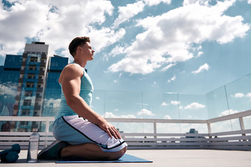 Tranquil young man is relaxing after training with dumbbells. He is sitting on knees and closing eyes for refreshment. Copy space in right side