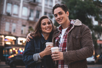 Waist up portrait of content couple bonding to each other outdoors. They are standing with coffee cups in hands and smiling