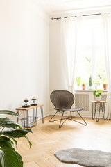 White background wall with empty space in a high ceiling room interior with a rattan and metal chair in the corner and side tables with candles and plants on a wooden herringbone flooring. Real photo
