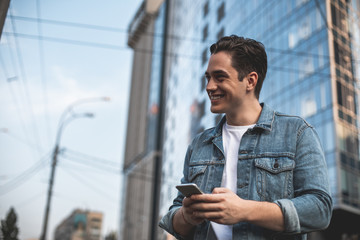 Side view of smiling male standing and holding smartphone in hands. He is writing message while waiting for girlfriend with pleasure. Copy space in left side