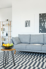 Grey sofa and table on patterned carpet in bright living room interior with posters. Real photo