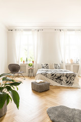 Comfortable bed with black and white cover and pillows, a rattan chair and a pouf standing on a wood parquet in a bright bedroom interior of a residence. Real photo