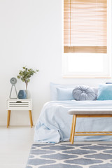 Window with wooden blinds in white bedroom interior with bed with light blue bedclothes, carpet...
