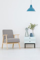 Patterned armchair standing next to small cupboard with clock and plant in vase in white room interior with blue lamp