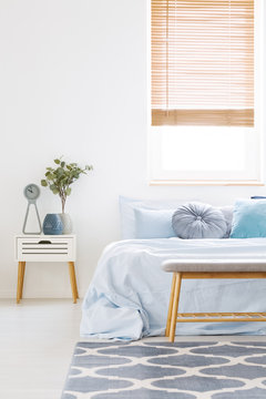 Window with wooden blinds in white bedroom interior with bed with light blue bedclothes, carpet with pattern and bedside table with clock and plant