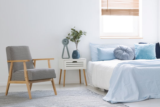 Armchair standing on striped carpet in white bedroom interior with bed with blue sheets and many pillows, wooden bedside table and window with blinds
