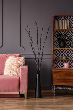 Creative black sculpture between a cozy, pink sofa with a furry pillow and an elegant, wooden bookcase in a dark gray living room interior