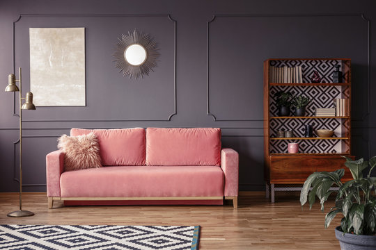 A marble beige painting and a sunburst golden mirror on a gray wall with molding in a stylish living room interior with a velvet, powder pink sofa and retro furniture