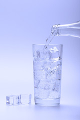 Pouring fresh water into glass with ice from a bottle. Ice cubes next to glass.