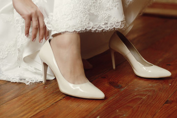 Close-up of beautiful bride in vintage lace white wedding dress putting on stylish white shoes, morning wedding preparation, wooden floor background