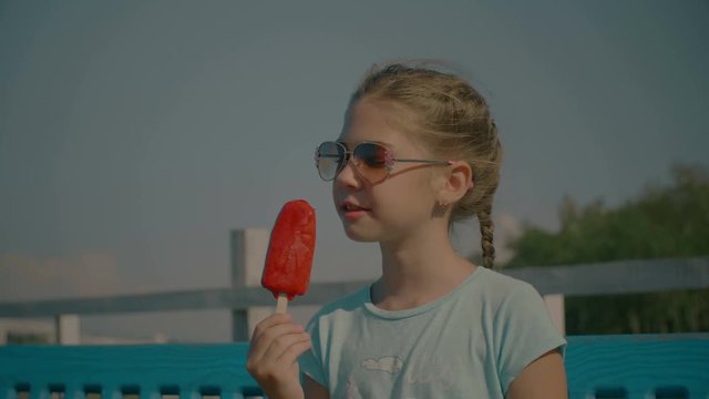 Portrait of lovely happy preteen girl in sunglasses eating delicious red fruit ice cream on stick in summertime while enjoying free time in park. Beautiful child eating popsicle outdoors. Slo mo.