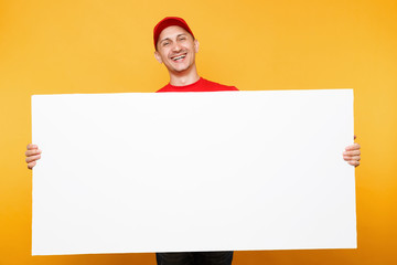 Delivery man in red uniform isolated on yellow orange background. Male employee courier in cap, t-shirt holding big white empty blank billboard with copy space, place for text image. Service concept.