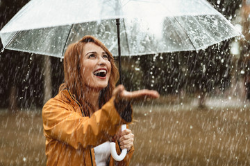 Waist up portrait of smiling girl standing outdoors with umbrella in hands. She is stretching hand...