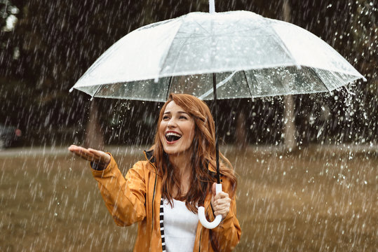 Waist up portrait of delighted girl standing outside and hiding under umbrella. She is stretching hand catching water drops with joy