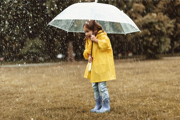 Full length side view of joyful small girl spending time outdoors. She is standing and holding umbrella hiding from water drops with sincere excitement