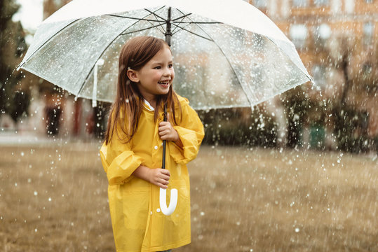 Side view of happy small girl standing on grass and holding umbrella. She is wearing raincoat and looking sideways with joy