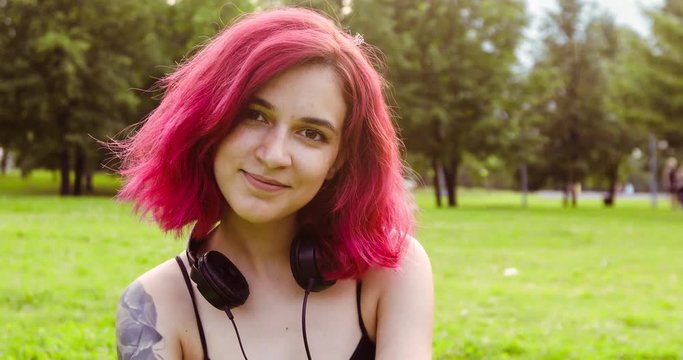 Young woman with hipster with burgundy hair looking at camera with headphones sitting in city park on grass