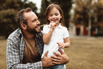Side view of delighted man squatting near small kid and holding her. Little girl is standing with...
