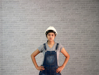 Technician woman ware white helmet with grey T-shirt and denim jeans apron dress standing akimbo on grey brick pattern background.