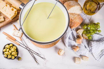 Gourmet Swiss fondue in traditional fondue pot, with forks, various cheeses, olives, bread and grape, white marble background copy space