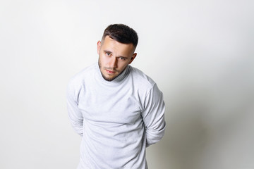 Fototapeta premium Close-up portrait of a sad man. Resentment of a man in white turtleneck his hands behind his back and lowered his head on grey background copy space