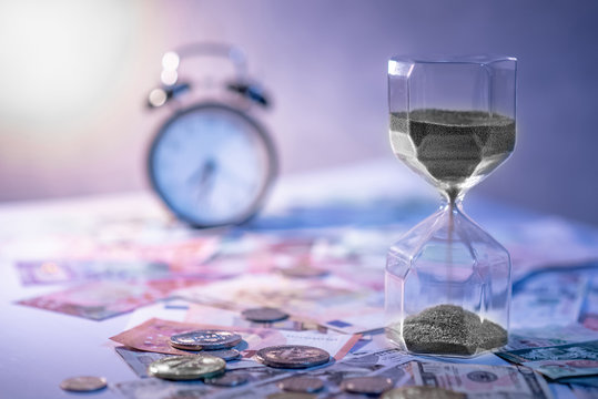 Sand running through the shape of hourglass on table with banknotes and coins of international currency. Time investment and retirement saving. Urgency countdown timer for business deadline concept
