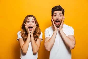 Deurstickers Image of young people man and woman in basic clothing screaming in surprise or delight and touching cheeks, isolated over yellow background © Drobot Dean