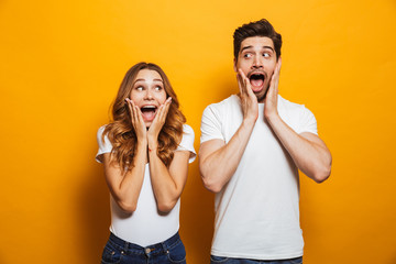 Photo of amazed couple man and woman in basic clothing screaming in surprise or delight and touching cheeks, isolated over yellow background - obrazy, fototapety, plakaty
