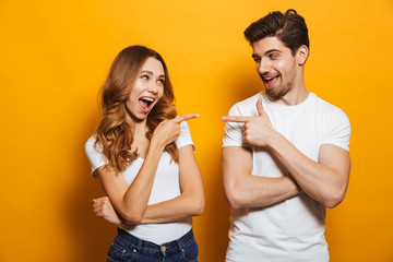 Image of happy young people man and woman in basic clothing laughing and pointing fingers at each other, isolated over yellow background