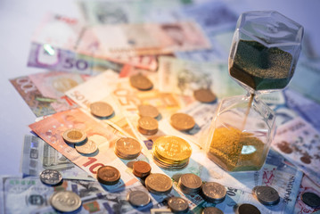 Sand running through the shape of hourglass on table with banknotes and coins of international currency. Time investment and retirement saving. Urgency countdown timer for business deadline concept