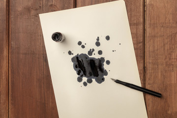 An overhead photo of an ink well with drops of ink and a nib pen, with copy space, on a dark rustic background