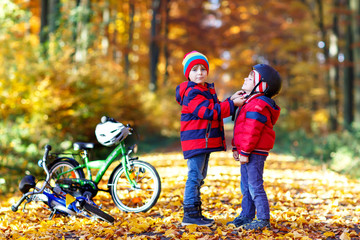 Two little kid boys with bicycles in autumn forest putting helmets