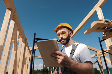 The man is a builder on the background of the roof of a frame house, in a yellow helmet and gray overalls. Uses a tablet