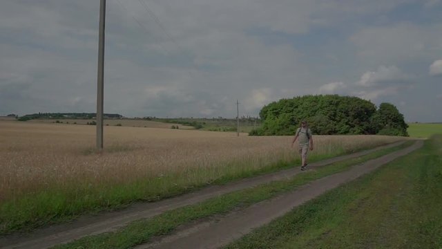 A man walking on a rural road. Adult. Against ripe wheat field. Stop to rest. The traveler with a backpack.