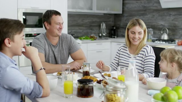 Joyous Caucasian family having breakfast together at home: mother, father son and little daughter talking and smiling while enjoying meal at kitchen table
