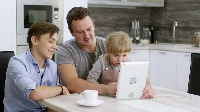 Happy dad sitting with cute little daughter and teenage son at kitchen table, smiling and laughing while girl making faces at camera of digital tablet