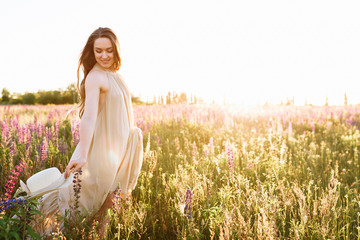 Beautiful dark-haired woman in a summer dress in a field of blooming lupine flowers with a straw hat in hands