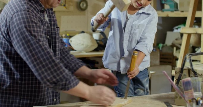 Tilt down of Asian male carpenter teaching little son how to use chisel and hammer on wood board while spending time together in workshop