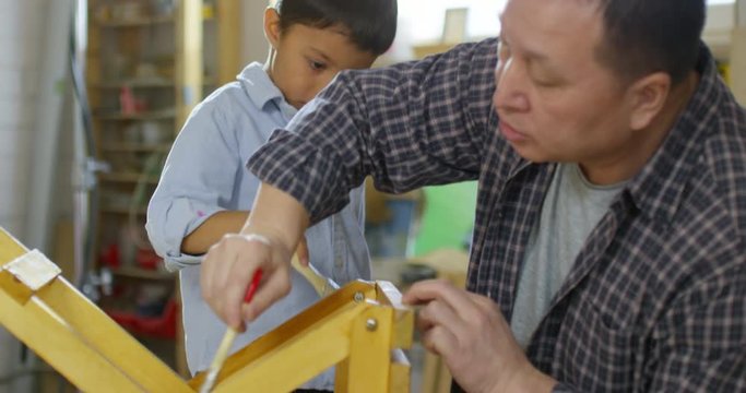 Handheld shot of happy Asian boy helping father staining wooden stool in garage