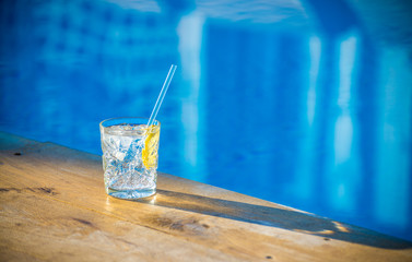 Glass of mineral water with lemon and ice near waterpool on the wood texture