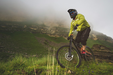 Fototapeta premium An adult mtb cyclist on a mountain bike at the foot of a cliff surrounded by green grass. Low clouds. North Caucasus. Russia